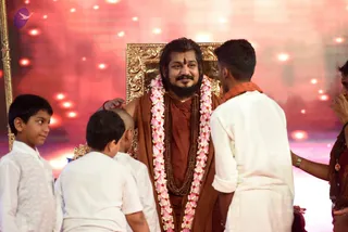 A seated man in a rust-brown robe and rudraksha mala sits on a gilded throne with a pink and white flower garland draped over his shoulders, with a red tilak on his forehead and a gold chain at his neck. A man in white clothes with an orange shawl stands on the right and rests a hand on his arm; several boys in white kurtas stand or crouch in the foreground, and a shaved-headed boy kneels nearby. The stage is backed by a pink-red lit wall with bokeh light spots. Swamiji (probable): the seated man wears long swept-back hair, a rudraksha mala and a gold necklace on a rust-saffron robe, and sits on a gilded throne as the central figure being honoured.