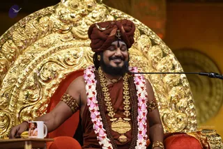 A smiling man with closed eyes sits on a red cushioned seat in front of an ornate gold carved throne back, speaking into a microphone on a boom stand. He wears a maroon-brown turban with a gold crescent and trident emblem, a maroon-brown robe, multiple strands of dark rudraksha beads, gold ceremonial necklaces with a large gold pendant, gold armbands, and long pink-and-white flower garlands. A white mug with a printed portrait sits on a small table beside him. Swamiji (probable): long-bearded figure in saffron-brown ceremonial robes, turban and rudraksha malas, seated on a gold throne as the speaker at a discourse.