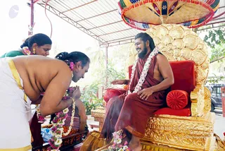 A photograph of a ceremony under a corrugated canopy outdoors, with green foliage behind. A seated man with long dark hair, a full beard, rudraksha-style garlands of white and pink flowers, and a rust-red robe smiles from an ornate gold throne with a fan-shaped gold back and red cushions, under a multicoloured fringed canopy. Two devotees in white dhoti with gold borders bow forward with folded hands, one wearing a green blouse behind them and a gold-bordered cloth. Marigold and pink flower garlands drape the seat and a wooden stand with offerings. The seated man is a person by appearance cues: long swept-back hair, ornate throne, and a presentation consistent with the page's context; the page does not state he is present, so this is set to unsure. Three people are visible.