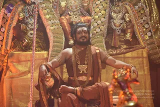 A photograph of a man seated on an ornate golden throne in front of a large, richly decorated backdrop of gilded deity figures with jeweled crowns, necklaces and ornaments. The seated man wears a rust-orange and brown draped cloth, a heavy gold necklace with a large gold pendant, multiple gold chains, gold armbands and bangles, and a gold crown. He has a red tilak mark on his forehead and long dark hair, and holds a golden bowl-like object in one hand. A tall striped staff or trident-like rod stands to the left, and blurred orange and gold objects appear in the foreground. Appearance cues match the page's Swamiji reference (long hair, crown, saffron-toned clothing, ceremonial gold necklaces), so this is a probable Swamiji in person seated on a throne as a central figure.