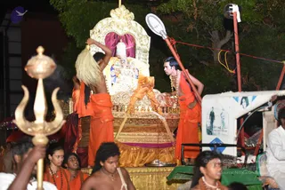 Night procession scene with a golden ornate ratha or palanquin carrying a deity murti dressed in white and blue with garlands, under a gold canopy with a red-maroon cloth backdrop. Two shirtless men in saffron-orange dhotis stand on the chariot tending the deity: one holds a large white chamara fan, the other, wearing a rudraksha mala and ceremonial beaded necklace, leans toward the murti. A golden trident (trishul) stands in the foreground beside a kalasha-topped staff. Women in orange saris and men in white and saffron clothing walk along the street, a red-and-white strip of paper-printed photos and a bright white light fixture visible on the right.