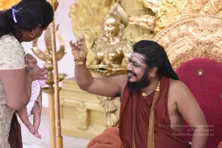A photograph of a long-haired bearded man in a rust-red sannyasi wrap and gold necklace, seated on an ornate gold-and-red throne, smiling and raising a gold-ringed hand toward a baby held by a woman in a cream patterned blouse at left. Behind him is a golden deity murti and a gold trident-topped pole with a kalasha-like lamp stand. A white vibhuti line marks his forehead. Swamiji (probable): long swept-back hair, rudraksha-style gold ornaments, ochre sannyasi clothing and a crown-like throne setting; the appearance cues qualify, so he is the probable Swamiji. The woman holding the infant is a devotee, and the baby wears a white outfit with pink accents.