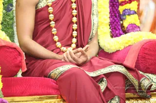 A seated figure in a maroon-red silk garment with a gold-bordered edge sits on a red velvet cushioned throne decorated with yellow and purple flower garlands. He wears a long necklace of large gold beads and holds his hands together in front of his lap in a meditative mudra with the fingers joined. Only the torso, arms and lower body are visible, with the face cropped out of frame. The setting suggests a satsang or public talk with ceremonial flower decoration.