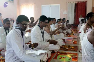 A photograph of a ritual scene in a plain room with pink walls and a red curtain. Several men in white dhotis and white shirts sit cross-legged on mats in rows, many wearing dark lanyard-style cords with rudraksha-like beads. They hold brass bells and incense sticks, with smoke drifting through the air, and open books or papers rest on their laps. In front of them are brass plates with flowers, turmeric-yellow powder, oil lamps lit on the floor, and small bowls of food offerings. Standing behind them at the far right is another figure in white. Nobody's face is singled out for identification here; the scene shows a group pooja with bells, incense and lamps.