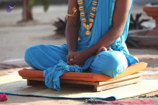 A person in a blue sari sits cross-legged on an orange cushion placed on a low wooden platform, resting on a woven mat. Only the lower body, arms and hands are visible; the face is cropped out. The figure wears a gold multi-bead necklace with a bird-shaped pendant and a gold armlet shaped like a serpent on the upper arm. The hands are clasped in the lap, and a corner of the blue cloth is draped over the wooden seat. The background shows a paved courtyard, potted plants and a small pink flower at the lower left.