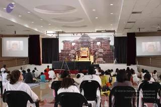 A large hall with a central stage where a framed portrait labelled as Paramahamsa Nithyananda sits on a gold-trimmed throne-like platform decorated with red flowers, flanked by two golden lamp stands. Behind the stage is a large backdrop of a stone temple with carved lion figures. Two projection screens on either side show the same portrait. Around thirty devotees sit on the floor and on black plastic chairs, most wearing white garments, with a few in orange and green. The audience faces the stage during a satsang gathering.