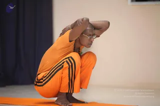 A photograph of a shaved-headed man performing a deep squat yoga posture on an orange mat in a plain room with a cream wall and a dark blue curtain at left. He wears an orange short-sleeved top, orange trousers with black side stripes, wire-framed glasses, a red bindi, and a dark rudraksha mala around his neck. His hands are clasped behind his head, elbows wide, with bare feet on the mat. a person candidate appearance is ambiguous given only orange sportswear and no crown, long hair, or ceremonial gold necklace, so identity is uncertain.