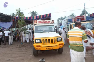 A white and yellow utility van with a red and white banner reading நித்யானந்தா stands on a dirt road in a village relief camp. Striped awning in red, green and blue shades overhangs the scene. Several men in white dhotis and shirts, some wearing caps and one with a grey hard hat, move about. A man in a green and yellow striped shirt stands in the foreground on the right.