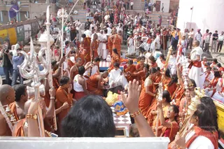 Outdoor photograph at a riverside ghat in Varanasi, with a crowd of devotees on stone steps behind. In the foreground, several people hold tall silver staffs topped with trident (trishul) finials and small bells. Many men and women wear saffron-orange robes, and some wear white dhotis and white shawls. A seated figure in white garments sits at the centre on a checkered red-and-white platform, with saffron-clad devotees around, some with hands raised in greeting or devotion. Many people are in the crowd behind, including onlookers in street clothes taking photographs.
