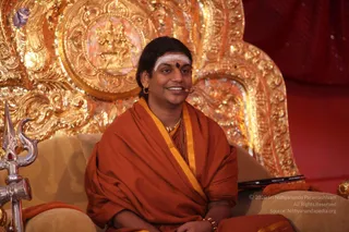 A smiling man sits on a gold-ornate throne-like seat with carved gilt backrest, a silver trident leaning at left. He wears an orange saffron shawl with a yellow border over an orange garment, a rudraksha mala, gold necklace and gold hoop earring, with white vibhuti horizontal stripe and red kumkum bindi on his forehead, and a small headset microphone near his face. Behind him a red curtain and a gold deity relief appear on the backrest. A small devotee-style figure is not visible; only one person is shown. a person cues include the saffron shawl, rudraksha mala, ceremonial gold ornaments and vibhuti, seated on a ceremonial throne during a discourse setting.
