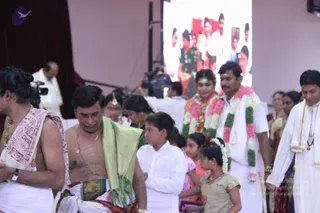 A photograph of a wedding-related ceremony in a hall with a red curtain and a large screen behind the stage showing a group photo. A bare-chested man in the centre wears a green shawl draped over his shoulder, a gold chain, and a dhoti with a dark-green and gold border, walking with a young boy in white and several children. On the right, a bride with a gold-and-red head ornament and a groom in a white dhoti and shirt, both wearing thick garlands of pink and white flowers with green leaves, walk with a man in white. A man at left has his hair in a topknot, wears a red-and-white shawl and green beaded necklaces. A cameraman and other attendees stand in the background.