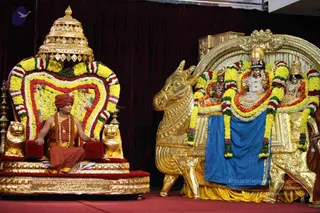 A photograph of a ceremonial stage with a seated figure on a golden throne (peetha) decorated with yellow and red flower garlands and a golden canopy with lion figures at its base. The seated person wears a red-orange turban, orange-saffron robes, a rudraksha mala and a gold necklace, with hands resting on the armrest. Beside the throne stands a gold-plated Nandi bull and a deity murti of a seated deity with a crown, ornate jewelry and blue cloth draped below, garlanded with yellow and red flowers. A dark red curtain forms the backdrop and a red carpet covers the floor.