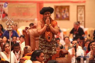 A photograph of a seated audience at a satsang-style event. In the centre, a person with swept-back dark hair, an orange and yellow sash, a leopard-print wrap, layered rudraksha-style orange malas and gold bangles stands beside a brown leather throne-like chair, palms pressed together in namaste with eyes closed. A tall silver trident stands at the left edge, and a microphone stand is at right. Behind them, many seated devotees in white, orange and blue clothing fill the hall; a banner and framed deity paintings decorate the back wall.