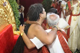 A photograph of a long-haired man in saffron-red robes and a rudraksha mala embracing an older, white-haired man in a white dhoti and red-bordered shawl, his head bowed close to the man's head. The long-haired man wears a gold wristwatch and bangles on his arm. The setting includes an ornate gold-carved throne with red upholstery at left and green foliage behind. In the background, women in white and gold-bordered saris and red blouses stand near a red curtain, one wearing a long gold necklace and red garland. Two people visible in the foreground are physically present, with additional people partially visible in the background.