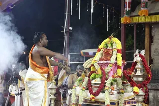 A night-time ritual at an outdoor temple-style pavilion. A bare-chested priest in white dhoti with an orange-and-gold shawl waves a ceremonial fan or chamara over a decorated structure garlanded with yellow and white flowers and red roses, topped with a pink element and offerings. Smoke drifts from the left. Behind, another white-clad man in a rudraksha mala and a bearded man stand near the ritual, with cameras on tripods at the edge. Brass oil lamps and a hanging garland border the scene, with striped and yellow decorations overhead. The ritual appears to be a puja or abhishekam; no deity form is clearly visible.