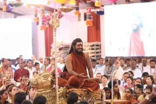 A seated man in a saffron-orange robe with long dark hair and a full beard sits cross-legged on a golden Nandi-shaped seat draped with a tiger-striped cloth, one hand raised in a gesture while smiling. A golden trident stands beside him. Behind him a large screen shows a figure in red robes. Dense crowds of seated devotees fill the hall, many in white and red clothing, some wearing rudraksha malas. Colourful paper lanterns and tassels hang from the ceiling.