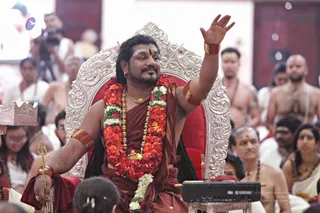 A man seated on an ornate silver-and-red throne raises his right arm in blessing while holding a small trident in his left hand. He wears a red-brown ochre robe, a dense garland of orange and white flowers, gold necklaces, gold bangles and a red-gold armband. His long dark hair falls past his shoulders and he has a tilak on his forehead. Behind him, seated devotees in white and cream clothing and bare-chested men with sacred ash markings watch the satsang. Some people in the background hold cameras. A Yamaha keyboard sits in front of the throne, and a silver umbrella-style canopy is visible at the left edge.