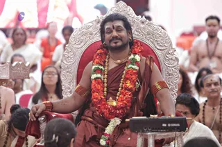 A man with long dark hair and a red bindi sits on an ornate silver throne with a red cushion. He wears a rust-red wrap, a thick gold necklace with a gold pendant, gold bangles and a long garland of red and orange flowers with white blossoms. A keyboard with a Yamaha logo sits on a small stand beside him. Behind and around him, seated devotees in white and orange clothing, one bare-chested man in the background wearing rudraksha malas, and a woman in an orange sari watch the gathering.