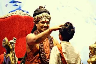 Photograph of a seated man on a red velvet throne with an ornate gold-trimmed back, wearing a leopard-print turban with a gold crown ornament, a red tilak on his forehead, long dark dreadlocks, a large rudraksha mala and an orange-saffron cloth draped over one shoulder. He smiles while reaching out to touch the head of a young boy in white kurta with an orange sash who stands before him. Beside the throne a gold-toned statue of a bull with a trident and a gold deity murti on the right are visible against a pale blue backdrop.
