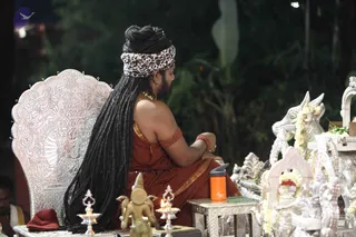 A photograph of a seated figure on an ornate silver throne with carved floral and figural back panels. The figure has very long dreadlocked hair falling down the back, a leopard-print headwrap, a gold necklace, and a rust-orange robe draped over the bare shoulder, with red thread bands on the upper arm and wrist. Beside the throne stand silver peetha-style ritual objects, a silver bull figure with yellow flower garlands, a small brass deity figure, two brass oil lamps with lit flames, a red cloth, and an orange water bottle on a silver side table. A partially visible man in white sits at the far left edge. The setting is a dark outdoor evening background with foliage. a person appearance is probable from the long locks, orange robe, gold necklace and throne, but the face is turned away and identity is uncertain.