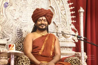 A seated man in a rust-orange turban and rust-orange draped robe with gold-edged borders sits on an ornate silver throne with intricate repoussé floral and scroll carvings. He wears a gold necklace and a red tilak on his forehead, and a microphone stand is positioned beside him. Behind him hangs a red curtain. A small framed picture sits on a silver side table at left, and a leopard-print cushion covers the seat. Swamiji (probable): long dark hair, rust turban and robe with gold border, and a gold necklace are visible cues, so this is a seated presenter of the satsang, with the throne.