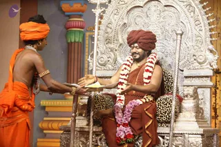 A seated man in a dark red-brown robe and matching turban sits on an ornate silver throne carved with floral and scrollwork, with leopard-print cushions. He wears long garlands of red and white flowers and a purple flower garland at his waist, and a small bindi on his forehead. In front of him a bare-chested attendant in saffron-orange cloth and turban, with gold armlets and a white beaded necklace, presents a tray with papers or offerings. Silver staffs stand on either side of the throne and a silver lion figure sits at its base. The backdrop is a painted stage set with orange and green pillars. This appears to be a stage-set discourse or ceremonial scene. The seated man is a candidate for a person, based on the throne setting, garlands and red-brown robe, though his appearance is only partially distinctive.
