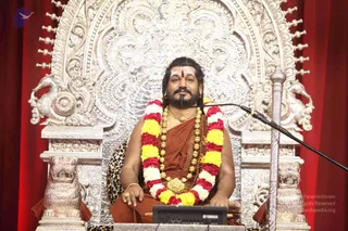 A seated man in saffron-orange robes sits on an ornate silver throne carved with floral scrollwork and mythical birds, with lion-head armrests and a red curtain behind. He wears a red, yellow and white flower garland, layered gold necklaces, a rudraksha mala, and a red tilak on his forehead, with long swept-back hair. A leopard-skin cushion rests beneath him. A microphone stands at his right and a staff with a silver top rests beside the throne; a Yamaha keyboard sits in front. Swamiji (probable) is seated in front, with long swept-back hair, a saffron robe, gold necklaces and a rudraksha mala, on a ceremonial throne, indicating the Paramahamsa Nithyananda figure at a satsang.