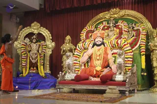 A photograph of a temple-style shrine room with a standing ornamented deity murti on the left, dressed in gold jewellery and a garland of yellow and purple flowers, seated on a blue cloth. In the centre, a large life-size cutout or printed image of a bearded man in an orange turban and orange robe, wearing a gold necklace and garlands, sits on a silver throne with a red cushion, flanked by silver lion figures and a multicoloured flower garland arch behind. On the left, a man in orange robes with bare upper body and white markings on his forehead and arms raises his hand, apparently waving incense or a lamp near the deity. A golden standing figure stands at the right edge. Oil lamp and flowers are visible on the floor. Because the central figure appears on a flat printed or cut-out image rather than as a physical person, it is counted as a depiction.