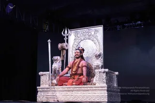 A bare-chested man with long dark hair sits cross-legged on an ornate silver throne carved with lions and floral panels, against a dark stage backdrop. He wears a rudraksha mala and layered ceremonial necklaces, orange-red dhoti cloth, bangles, and a leopard-print cloth draped behind him. A silver trishula stands at the right side of the throne, with a damaru drum hanging beside it and a silver staff beside the throne's left arm. Stage lights and truss are visible above. Swamiji (probable) is seated on the throne, with the qualifying cues of long hair, rudraksha mala, ceremonial gold necklaces and orange-red clothing.