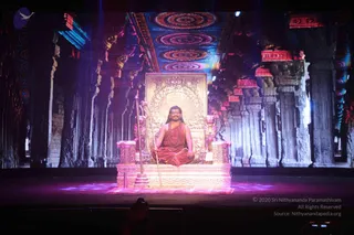 A seated man in orange-red robes sits cross-legged on an ornate golden throne on a stage, with lion figures flanking the seat and a golden staff standing beside it. He wears a rudraksha mala and a ceremonial necklace, with long hair and a calm expression. Behind him, a large projected temple hall with carved pillars, red and blue patterned ceiling medallions and illuminated backdrop fills the stage. Purple stage lighting falls on the floor. A small silhouetted figure sits in the lower foreground. Swamiji (probable): the seated figure in orange robes with long hair, rudraksha mala and ornate ceremonial necklace, seated on a throne.