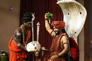 A seated man in a rust-red turban and rust-orange robes, wearing rudraksha malas and a gold-beaded necklace, raises a bunch of green leaves with one hand while smiling. Beside him, a younger attendant with long dreadlocked hair, white horizontal body markings, orange wrap and gold necklace holds a white cloth-wrapped kalasha decorated with purple and gold embroidery. Behind them stand a silver trident and a silver ceremonial staff, a red curtain and a large silver throne with a cobra-hood backrest set with red jewels. A small plate of offerings sits between them. Swamiji (probable): the seated turbaned figure, identified by his turban, rudraksha malas and ornate ceremonial robes on a throne.