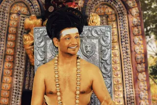 A bare-chested man with a tall black turban-style hair wrap and a white vertical tilak on his forehead stands in front of an ornate silver throne with carved lion-face motifs. He wears a long necklace of large gold beads and smiles toward his left. Behind him are gold and orange decorated arches, a gold sculpted figure, and foliage at the right edge. Appearance-based label: a person, with long hair wrapped in a tall turban, a gold bead mala and a bare chest as qualifying cues, shown in person.