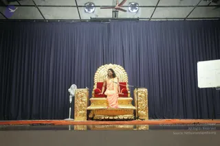 A photograph of a stage with a navy blue curtain backdrop and an ornate gold throne with red velvet cushions at centre. A seated figure in a peach-orange wrap with long dark swept hair and a beaded necklace sits on the throne, hands resting on the armrests. A white standing fan and a whiteboard with diagrams stand to the sides, and an orange carpet runs along the stage edge. Swamiji (probable): the figure has long swept-back hair, an orange wrap and a beaded necklace, seated on a gold throne, so the label is based on these appearance cues.