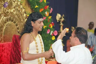 A seated man with long dark hair and a white tilak on his forehead wears a white dhoti-style garment, a gold ceremonial necklace of large gold beads, and a gold armlet. He sits on an ornate gold throne with red upholstery and clasps hands with a man in a white shirt who raises his palm in greeting. Behind them stand a golden oil lamp with a lit flame, a backdrop of colourful flowers including pink, orange, red and yellow blooms, and a bystander in a striped shirt. The swamiji cues here are long hair, gold necklace and a seated throne, so the seated figure is labelled Swamiji (probable).