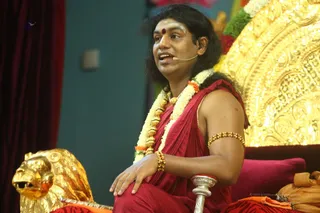 A man seated on an ornate golden throne with a carved golden lion armrest speaks into a headset microphone during a satsang. He wears a red-maroon draped garment over bare shoulders and chest, a long garland of white and orange flowers, gold bead bangles on his upper arm and wrists, and a red bindi on his forehead. His long dark hair falls past his shoulders. Behind him are a red velvet cushion and gold-tinted decorations, with a teal wall and a maroon curtain at the left edge. The seated speaker's appearance and setting fit the page's description of a satsang with a person, though the presence of a teaching is not visible in the frame. Shown as a single in-person speaker, identity uncertain from appearance alone.