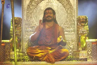 A seated man in saffron-orange robes sits cross-legged on a leopard-print cushion before an ornate gold throne flanked by two gilded lion sculptures. He wears a rudraksha mala and a gold necklace, has long swept-back hair and a red tilak on his forehead, and raises one hand in blessing. Ornate gold metalwork with carved figures forms the backdrop, with stage lighting and haze behind him. Probable Swamiji by appearance cues: long hair, saffron clothing, rudraksha mala and ceremonial gold necklace, seated on a throne.