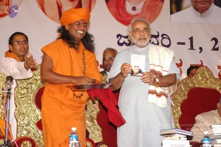 A stage event with two central men. A long-haired man in saffron-orange robes and a matching saffron turban holds a silver tray with a red cloth, smiling broadly, with a wooden rudraksha-style mala around his neck. Beside him a grey-haired, bearded man in a pale grey kurta with a cream and red-bordered shawl holds up a small printed booklet showing a photo of a smiling man. Ornate gold-and-red chairs, a microphone stand, water bottles and stacked books sit on the stage. A man in white with an orange scarf gestures to the left, and a seated man in the background wears glasses. Behind them a large banner shows Kannada text and portrait photos. The printed name card on the table reads Sri Narendra Modi, which attributes that name to the card, not to the man's appearance. A saffron-robed figure with long hair and a turban is a person by the appearance cues, presenting the tray and booklet.
