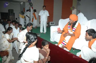 A photograph inside a hall with a green floor and white sofa-style chairs. A man in saffron-orange robes, an orange turban, a multicoloured flower garland and a red-and-white bead necklace sits on a white sofa, smiling and gesturing with his hands while speaking. Beside him on the right, a young man in white with an orange shawl sits on the sofa. Seated on the floor in front are a woman in a white sari with a long braid, a young girl in a brown checked dress, and a woman in white and gold near the left. Behind them, men in white dhotis and shirts stand and sit; one man in a white shirt stands at the back holding a pendant necklace. A small white air-conditioner unit and orange-and-white striped wall panels are visible, along with a red-draped low platform in the foreground.