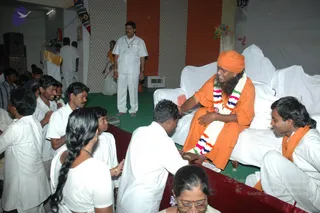 A photograph of an indoor devotional gathering with a green carpeted stage and white-draped sofas. A seated man in orange clothing, with an orange turban, a long white and multicolour garland, and long hair swept back, sits smiling on the right while a devotee in white kneels before him and touches his foot. Around them, several people in white ritual clothes sit or stand. A standing man in white with a pendant necklace watches. A woman in a white sari with a long plait stands at left, and a woman with glasses sits in the foreground. Orange and red striped drapes and a flower decoration hang on the back wall.