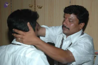 A photograph of two men in white shirts in an indoor room with pale wooden cupboards and brass handles behind them. The man on the right, with short dark hair, a moustache and a red tilak on his forehead, has his eyes closed and both hands placed on the head of the man seen from behind, in a blessing gesture. He wears a brown beaded necklace over his shirt. The seated figure on the left has short dark hair and a white collared shirt. Appearance is not distinctive enough to confirm the a person identity, so the figure is described by role as a man giving blessing.