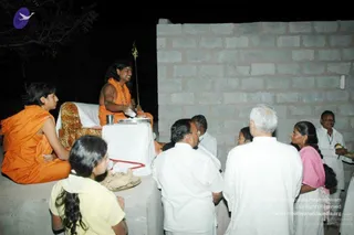 Night outdoor scene before a plain whitewashed concrete-block wall. On a raised white-draped platform sits a figure in saffron-orange robes with long dark hair and a bare upper body, holding a long golden staff and a small silver plate. Beside them, a second figure in orange robes with short hair sits on the platform. Several people in white clothing stand in front of the platform, including a man with white hair, a woman in a pink top, and a girl in a yellow top in the foreground. A man in white on the right holds a small object. Strings of cloth and a plate of offerings sit on the platform edge. A purple and white logo appears at the upper left.