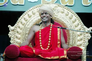 A smiling man in a cream turban with a red bindi sits cross-legged on an ornate gold throne with carved scrollwork and red velvet cushions. He wears a red shawl with gold border over a red dhoti-style garment, and a long gold bead necklace. A microphone on a stand is positioned beside him. Behind him is a green panel with large gold lettering.