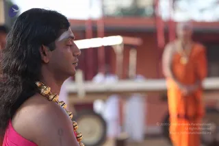 Close profile photo of a man with long dark hair falling past his shoulders, eyes closed, a white vertical mark on his forehead, wearing a pink sleeveless garment and a heavy gold bead necklace of large ornate beads. Behind him, out of focus, a second figure in orange robes and a gold pendant necklace stands with hands folded, beside indistinct white-clad people and red-orange buildings and a wooden structure. The foreground man appears to be in meditative stillness. a person cues visible on the foreground man: long swept-back hair, ornate gold necklace, and a white forehead mark; he is presented as a devotee in meditation position.