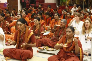 Photograph of a seated group of women in orange and rust-red saris and robes, many with rudraksha malas and white vibhuti lines on their foreheads, performing a ritual with small oil lamps and flower-filled brass plates. Several hold lit lamps or offering dishes; a woman in the foreground at right wears white with a mala and holds a lamp, and another in white at upper right also holds a lamp. The seated rows fill the hall, with more devotees behind and red cushioned seating at the back. Gold-toned ritual items and a metal tray with offerings sit on the floor in the front.