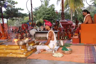 Outdoor ritual homa beside a fire pit with flames rising from a stepped wooden platform. A young man in white dhoti and shawl with a red turban sits cross-legged on an orange mat, tending the fire with brass vessels, a bell and flower-filled dishes nearby. A man on an orange-cushioned seat at right, wearing orange robes, rudraksha-style neck beads and a flower garland, sits with bare feet forward. Dozens of devotees in orange and red robes sit in rows under palm trees, with a red temple pillar and a camera operator behind.