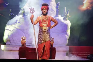 A man stands on a lit stage in front of a large white rock-like backdrop with carved deity figures and a trident-topped sculpture, with mist on the floor. He wears a red turban, a red sleeveless garment with a leopard-print wrap, a red dhoti with gold trim, a multi-strand rudraksha-style mala and a large gold pendant, gold armbands and bangles. He holds a tall twisted trident-headed staff in his right hand while his left hand rests on his hip. Golden lion figures flank a red velvet throne-like seat at his side. Stage lighting in purple and green fills the background.