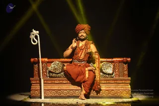 A seated man in saffron-orange ritual dress sits on an ornate carved wooden throne with leopard-print cushions, against a dark stage backdrop with green-yellow spotlight beams. He wears a red-orange turban, rudraksha mala necklaces, a gold chain, bangles and a sash, and raises his right hand in a blessing gesture. A silver serpent-headed staff stands at left and a patterned rug lies at his bare feet. Probable Swamiji by the page-described setting and visible cues of turban, rudraksha malas and saffron attire, seated on a throne in a presentation hall.