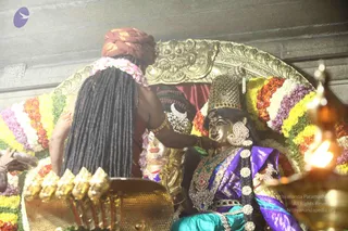 Photograph of a ceremonial scene in a temple-like hall. In the foreground, the back of a figure with long dark dreadlocked hair, a rust-red turban and a pink-and-white flower garland, wearing gold armlets and holding a gold ritual object with golden finials. Facing them is a golden-faced murti dressed in a purple and teal silk sari with jeweled ornaments, a tall jeweled crown, a crescent-moon ornament and a long braid decorated with white flowers, seated before a gold arched backdrop framed with orange, yellow, white and purple flower garlands. A lit brass oil lamp appears at the right edge. The figure in front appears to be a ritual attendant or devotee; no identifiable a person candidate is visible, and the scene is a deity murti setting.