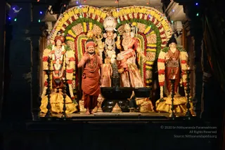 A photograph of a decorated shrine at night with a black Shivalinga on a stone yoni base at centre. Behind it stand larger deity figures of Shiva and Parvati, with the goddess in a pink and gold sari and a tall ornate crown, and the god holding a trident. Flanking the shrine are two standing figures in gold and red costume. In front of the linga, a man in a saffron-orange robe, red head-wrap and multiple rudraksha malas stands barefoot with one hand raised, a probable Swamiji by appearance (distinctive saffron clothing, red turban and rudraksha malas). Marigold and rose garlands frame the arch, with oil lamps on tall brass stands and string lights above.