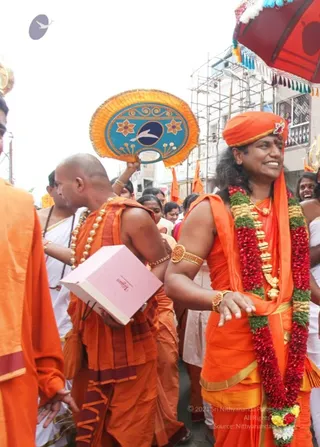 A street procession in a town, with a man in orange robes and an orange turban at the right, wearing a long red and white flower garland, gold necklaces and a gold armlet, smiling and gesturing with his hand. Beside him stands a bald man in orange robes holding a pink book, wearing a gold-beaded necklace and a rudraksha-style bead mala. Other orange-clad devotees and white-clad people walk behind, and a fan-shaped orange and blue ceremonial fan is held aloft. A red umbrella with tassels appears at the top right, and scaffolding and multi-storey buildings line the street.